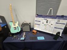 A breadboard setup of Omni-Pedal with an amplifier and guitar in the background, along with the Project Poster to provide additional context.