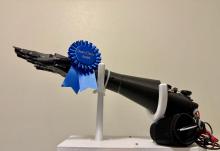 A side profile of a black 3D-printed prosthetic hand and forearm displayed on a white stand. A blue ribbon reading "Dean's Choice Award" is fastened around the wrist, and a black sensor cuff with wires rests near the base of the arm.