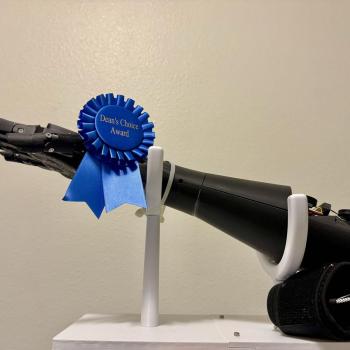 A side profile of a black 3D-printed prosthetic hand and forearm displayed on a white stand. A blue ribbon reading "Dean's Choice Award" is fastened around the wrist, and a black sensor cuff with wires rests near the base of the arm.